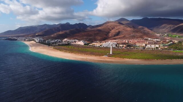 Aerial drone sweeps along Fuerteventura beach past dunes, small resorts, and a white lighthouse. Navy ocean meets turquoise shallows as clouds cast moving shadows.