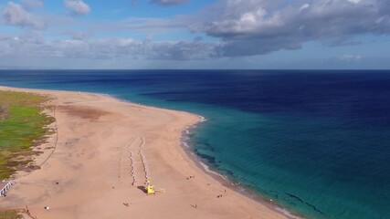 Aerial view of a sandy spit in Fuerteventura, Canary Islands. Turquoise shallows meet deep Atlantic blue. Lifeguard tower, sunbeds, cafe, swimmers, and walkers.