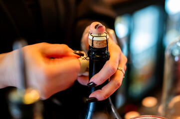 person's hands with dark nail polish opening a bottle of sparkling wine or champagne. The cork and wire cage are visible against a dark, blurred background