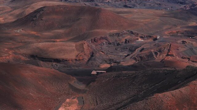 Aerial drone passes over eroded crater rims, lava hills, dirt tracks, fenced plots, and a small industrial site on Fuerteventura in afternoon light with long shadows.