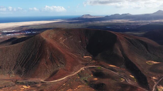Aerial view of a rust red crater, lava cones, dunes, and ocean on Fuerteventura, Canary Islands. Wind turbines, road, and small settlements appear in daylight.