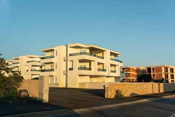 New modern white apartment complex with glass balconies and parking lot next to building under construction in Mediterranean town. Coastal real estate development, property investment, growth.