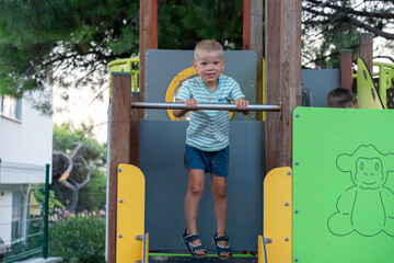 Happy blonde boy standing on playground platform holding metal bar above slide looking at camera on colorful play structure. Concept of confident childhood, outdoor fun, summer vacation, active kids.