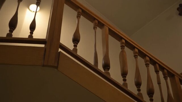 A staircase with ornate balusters is illuminated by a wall light in a dimly lit interior setting viewed from below.