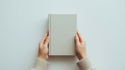 Person holding a blank light grey book with both hands, top view on a solid light blue background.