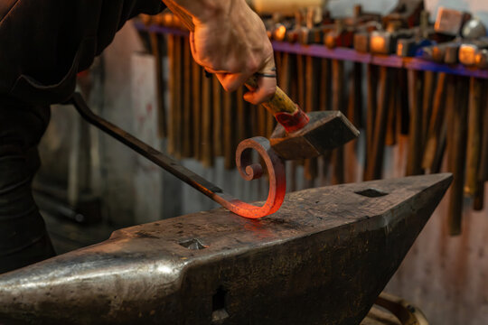 Gros plan sur un forgeron martelant une pi&egrave;ce de m&eacute;tal chauff&eacute;e sur une enclume, illustrant le travail artisanal de forge et la fabrication traditionnelle en ferronnerie.