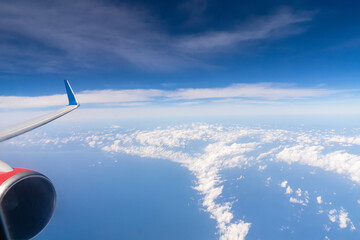 Obraz premium Clouds stretch across the sky as seen from an airplane window. A wing is visible on the right side, with the ocean below reflecting sunlight. This scene shows the vastness of the sky. High quality