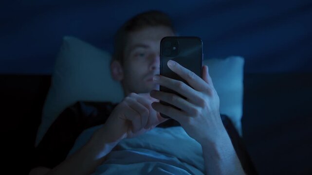 Young man lies in bed holding his mobile phone, scrolling social media at night with his face glowing under blue screen light as insomnia and digital distraction keep him awake. Screen addiction