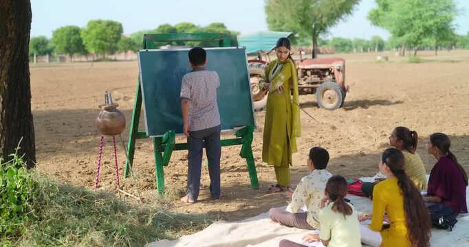 Indian happy young adult gen z female do work talk to villager girl boy sitting on mat rural area. teen male child writing study look board clap hand enjoy desi life open day class field ground place