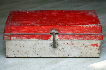 Old weathered metal storage trunk with a rustic latch and red paint. Visible rust patina, scratches, and worn texture on an antique security box. Close-up industrial container.