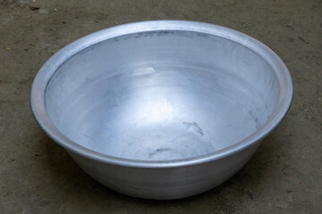 Large silver aluminum basin placed on a natural earth ground. Traditional domestic tool for washing clothes and daily chores in rural villages. Authentic household equipment and metalware.