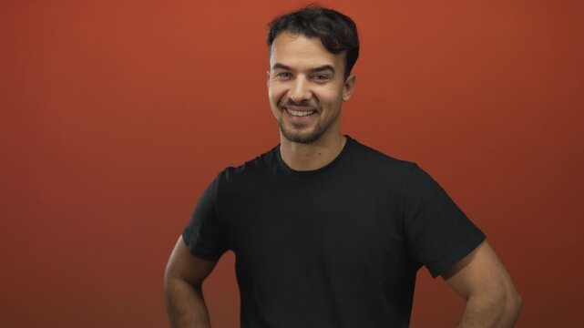 Young hispanic man smiling confidently in black shirt against vibrant red background, hands on hips radiating positivity and charm in professional portrait setting