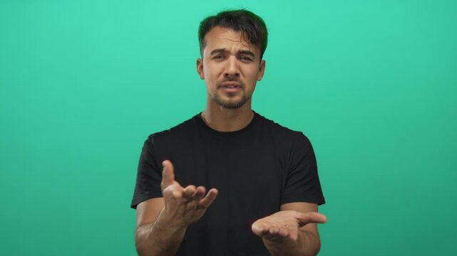 Young hispanic man gesturing for money over an isolated green background, emphasizing his expression and casual clothing in a studio setting