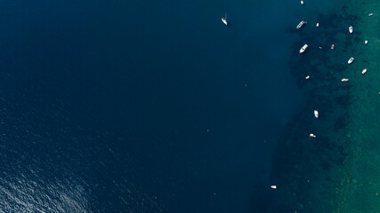 An aerial top-down view of a deep blue sea where several small white boats are anchored near a rocky coastline. The clear water reveals dark underwater formations and shallow turquoise areas.