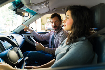 Driving instructor teaching adult woman student during a practical lesson inside a car, pointing and explaining traffic rules