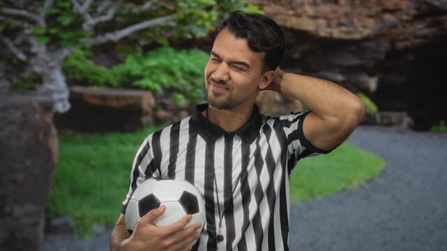 Young hispanic man referee with soccer ball in park outdoor setting, dressed in striped shirt, pensive expression, surrounded by greenery and natural environment.