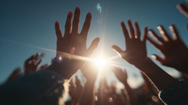 Diverse young adults' hands reaching up in celebration outdoors under a clear blue sky with dramatic golden hour backlighting and strong lens flare.