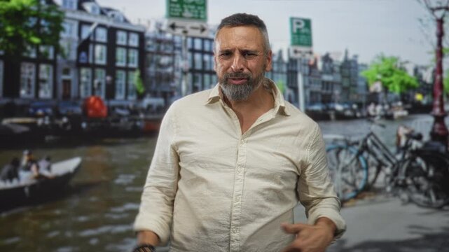 Man points finger at camera on street by canal in amsterdam, beard and rolled sleeves, bracelets visible; frustration confrontation.