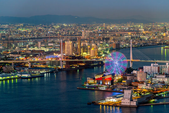 High angle night view of Osaka Bay with illuminated Tempozan Ferris Wheel and Minato Bridge. Vibrant cityscape of Osaka, Japan, at twilight with glowing urban lights and beautiful harbor skyline.