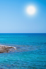 Vertical seascape with bright sun, clear blue water, and rocky coast, creating a calm summer atmosphere.