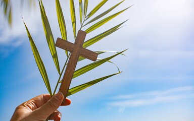 Palm sunday symbol with wooden cross on green palm leaves against blue sky with white clouds and sunlight with hand
