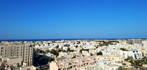 Two storey white buildings of Muscat city center and the Gulf of Oman with Al Fahal island in the distance seen from the rooftop. Panoramic city view