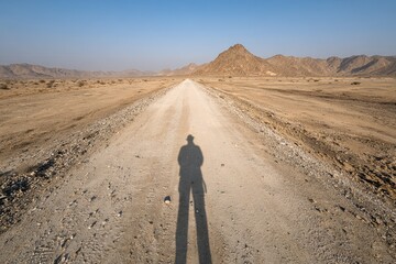 A lone traveler's shadow stretches along a dusty desert road towards distant mountains under a clear blue sky