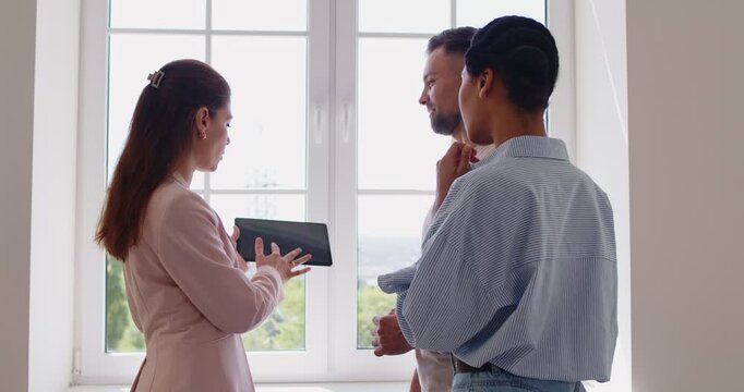 Real estate agent showing tablet screen with contract information to young couple on home tour. People standing by window, professional realtor talking to man and woman to explain details of deal