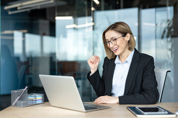 Fototapeta premium Young businesswoman triumphantly cheering and clenching her fist while looking at a laptop in a contemporary office setting, experiencing achievement and good news online