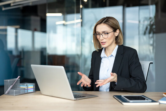 Businesswoman experiencing frustration and stress during a video call on her laptop in a modern office, communicating problems with hand gestures and a worried expression