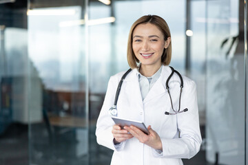 Smiling female doctor wearing a white lab coat and stethoscope, holding a digital tablet and looking at the camera, symbolizing medical technology and professional healthcare