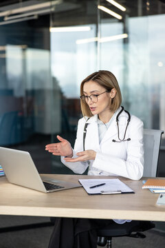 Female doctor in lab coat with stethoscope consulting patient via laptop video call from a modern office, providing telehealth services and remote medical advice