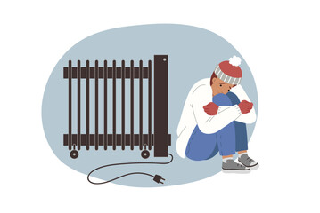 Man is freezing sitting near non-working electric radiator during power outage in house. Guy wrapped in winter clothes is located indoors and is freezing due to lack of heating in apartment.