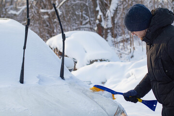 To clean the car from snow. A man in a jacket and hat cleans his car from the snow near the house. A man in a black jacket cleans snow from his car with a brush.