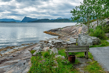 Benches are placed on a rocky shore overlooking calm water. Mountains are visible in the background under cloudy skies. Greenery is along the pathway beside the water.