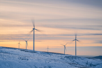 Multiple wind turbines stand in a snowy landscape as sun sets on a winters day