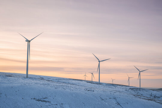 Multiple wind turbines stand in a snowy landscape as sun sets on a winters day - Powered by Adobe