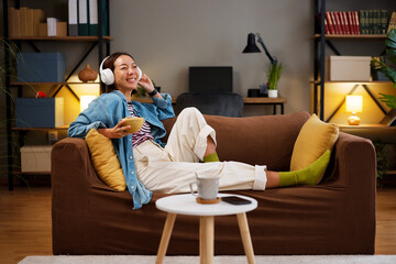 Cheerful Asian woman relaxing on sofa with headphones and a snack