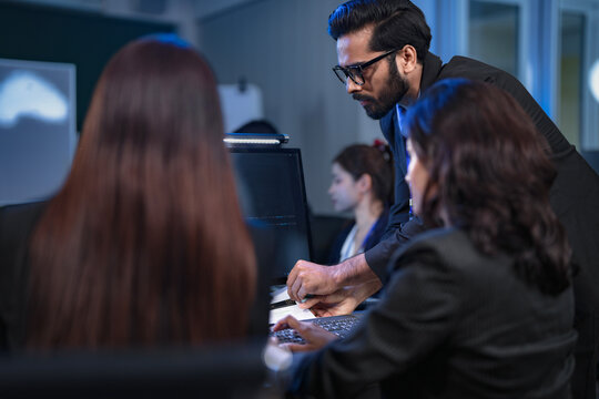 A senior data scientist with glasses points out an anomaly in a machine learning model's code to two female AI engineers, as they finalize the algorithm for a new predictive analytics service.