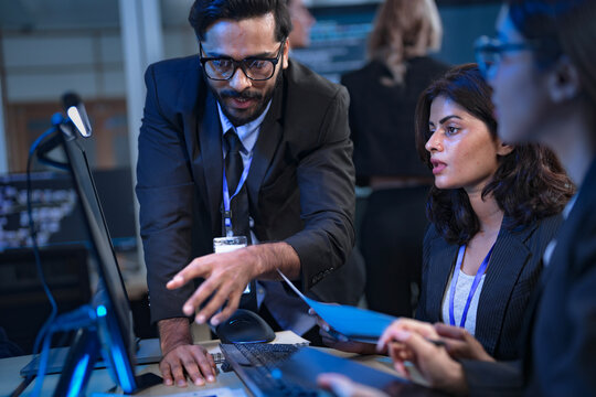 A senior data scientist with glasses points out an anomaly in a machine learning model's code to two female AI engineers, as they finalize the algorithm for a new predictive analytics service.