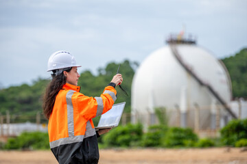 Female environmental scientist in safety gear standing and using laptop and radio for monitoring ecological impact inspection near industrial gas storage tank at fuel terminal site.