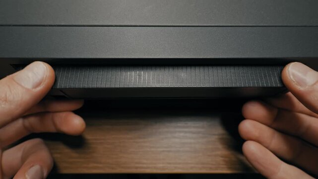 Male hands eject and pull out black VHS cassette tape from vintage VCR player slot. High angle view captures mechanics of retro analog video technology on wooden table.