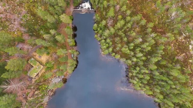 straight norway narrow forest reservoir bordered by dense pine trees small dam whitewater spillway footpaths rustic buildings sit among mossy woodland reveals from down drone view 