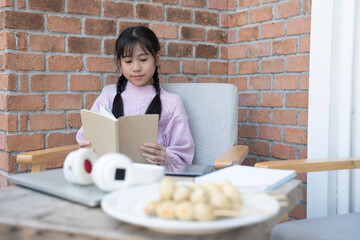 Young Asian girl reading a book at home. Smiling young elementary school girl holding and reading book sitting on a table at cozy home, education concept