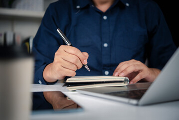 Close up businessman hand writing plan on notebook with pen. Freelancer man working on laptop, taking note of business agenda, study schedule, and checklist strategy in office.