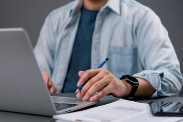 Close up creative designer man holding pencil and working on laptop computer. Freelancer student studying online course, planning business project with smartphone on desk in home office.