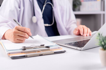 Close up doctor hand writing medical record on clipboard and laptop computer. Physician working on patient diagnosis report, healthcare insurance prescription and telemedicine in hospital office.