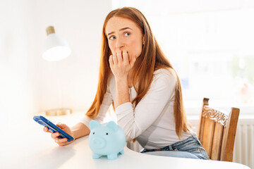 Young woman thinks about finances while holding a phone and sitting near a piggy bank