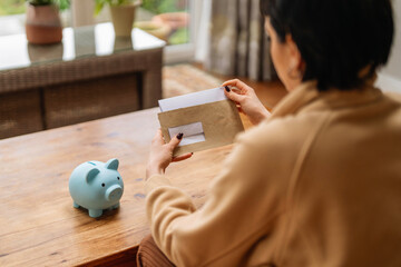 Person opens envelope near blue piggy bank on wooden table during day at home