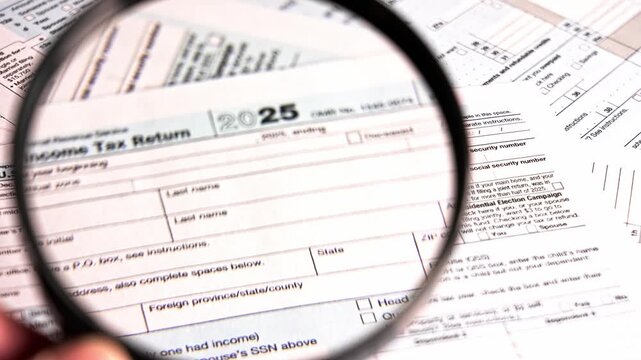 A person examines various tax documents and 1040 with with a magnifying glass. The focus is on details like numbers and sections relevant to tax filing. It takes place during tax season at home.